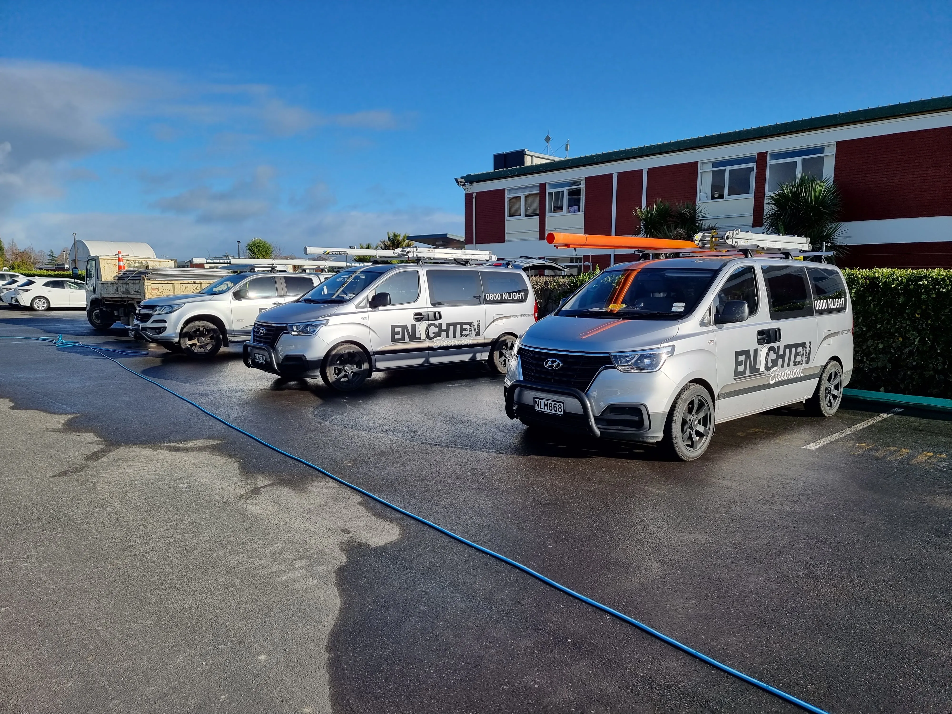 Three Enlighten Electrical branded vans and utes parked in a line in a yard after rain