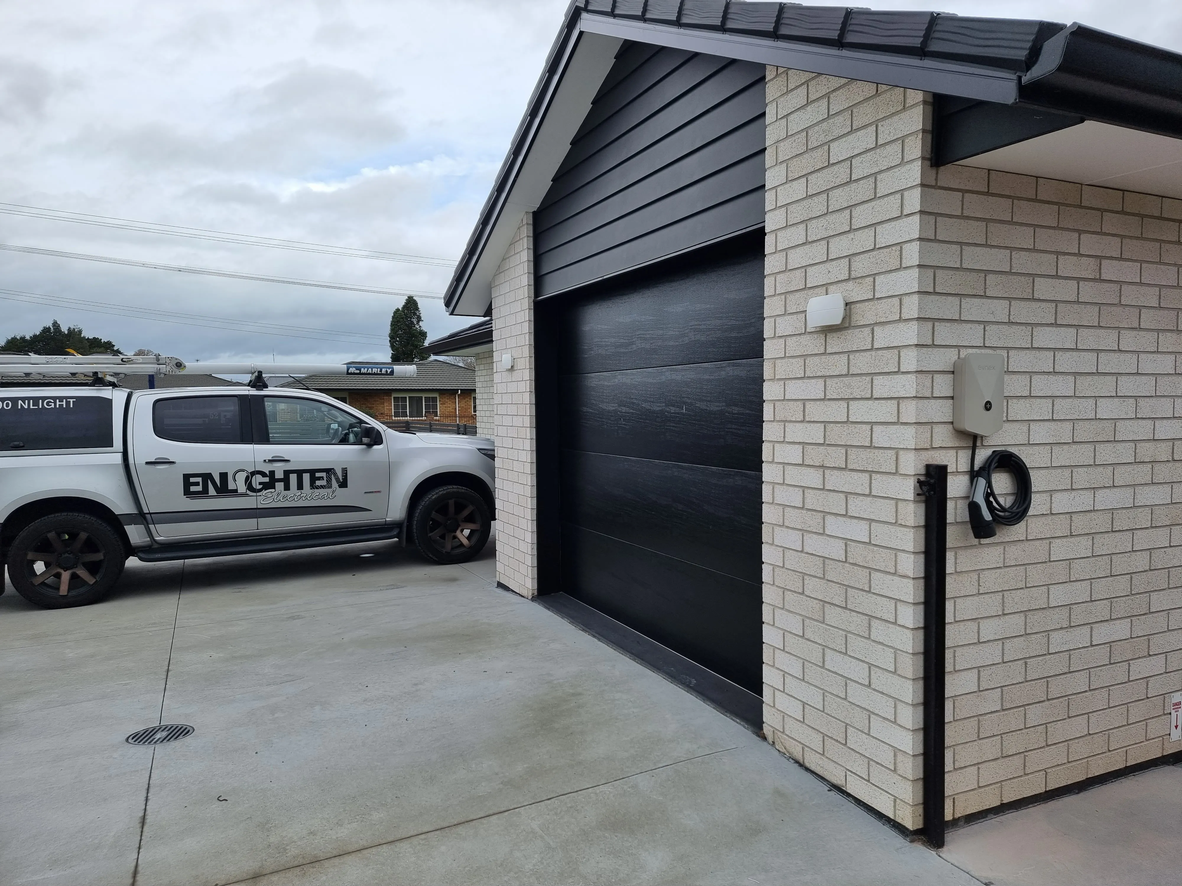 An Enlighten Electrical ute parked at a new brick house with a wall mounted EV charger beside the garage door